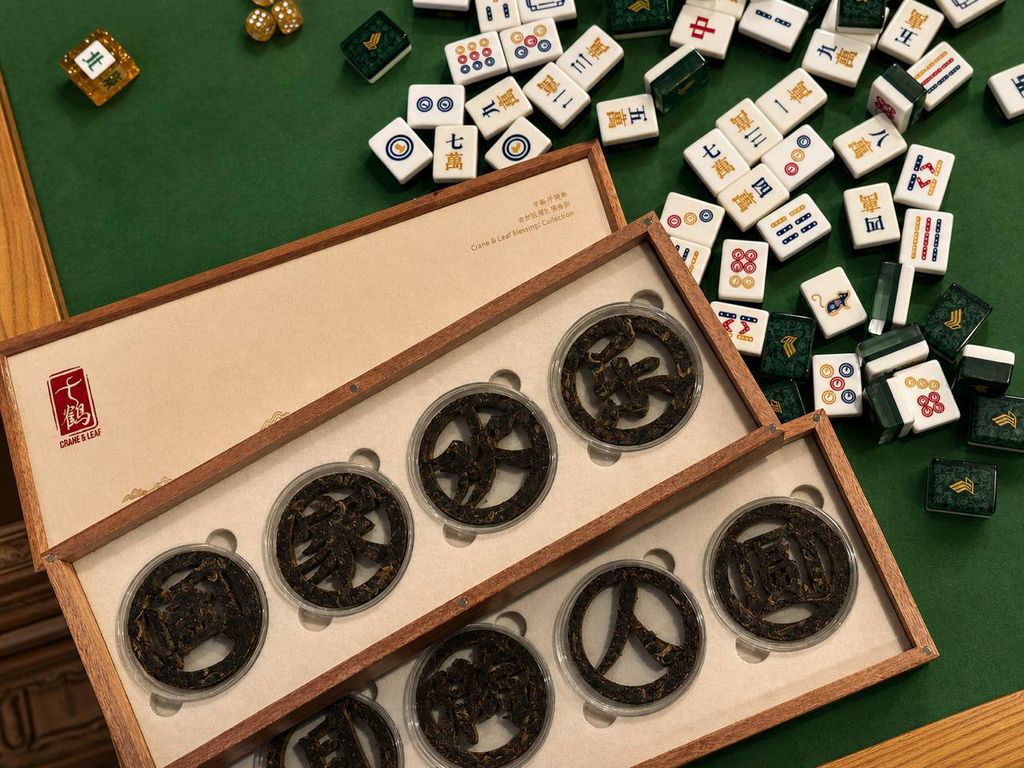 An open 千鶴 wooden tea gift box displaying eight carved pu’er discs on a green mahjong table.